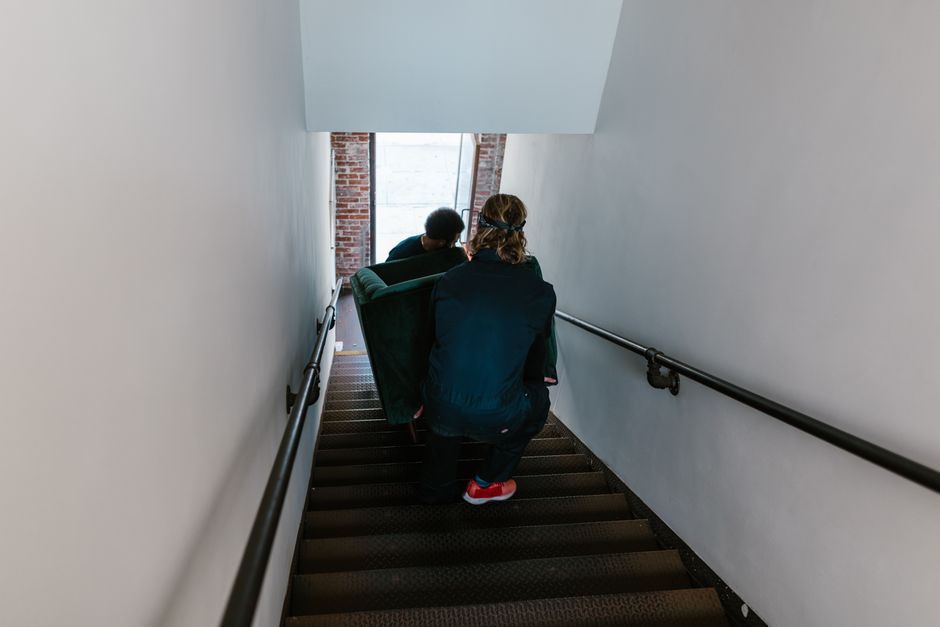 Two men carefully move a couch down a staircase in an indoor setting.